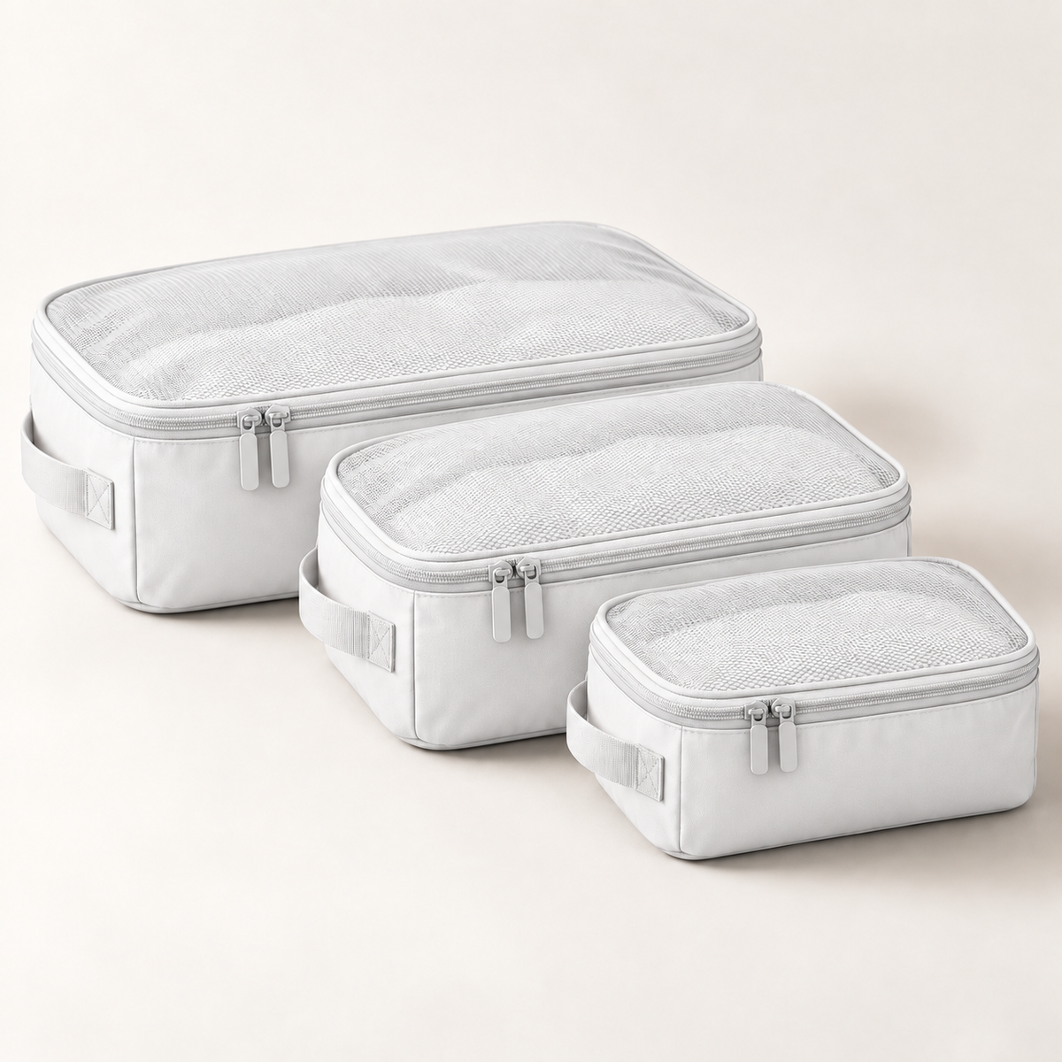 Three light gray packing cubes of different sizes arranged diagonally on a light beige background. Each cube features a zippered top with a mesh window, a side handle, and silver zipper pulls.